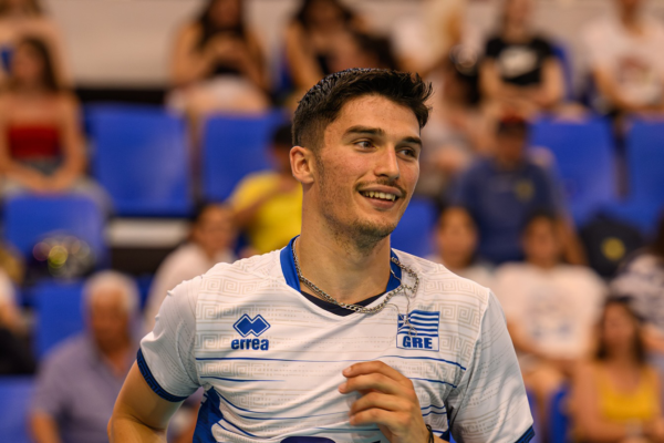 Smiling male athlete wearing a Greece jersey stands on a sports court with blue stadium seats in the background.