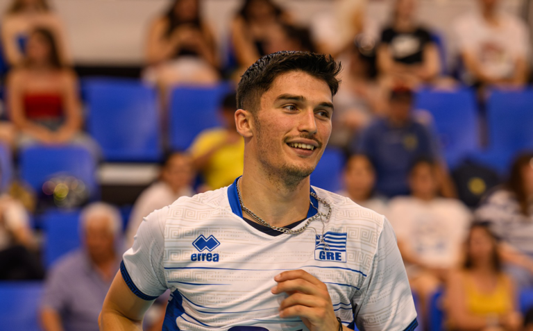 000 The Volley Lovers Smiling male athlete wearing a Greece jersey stands on a sports court with blue stadium seats in the background.
