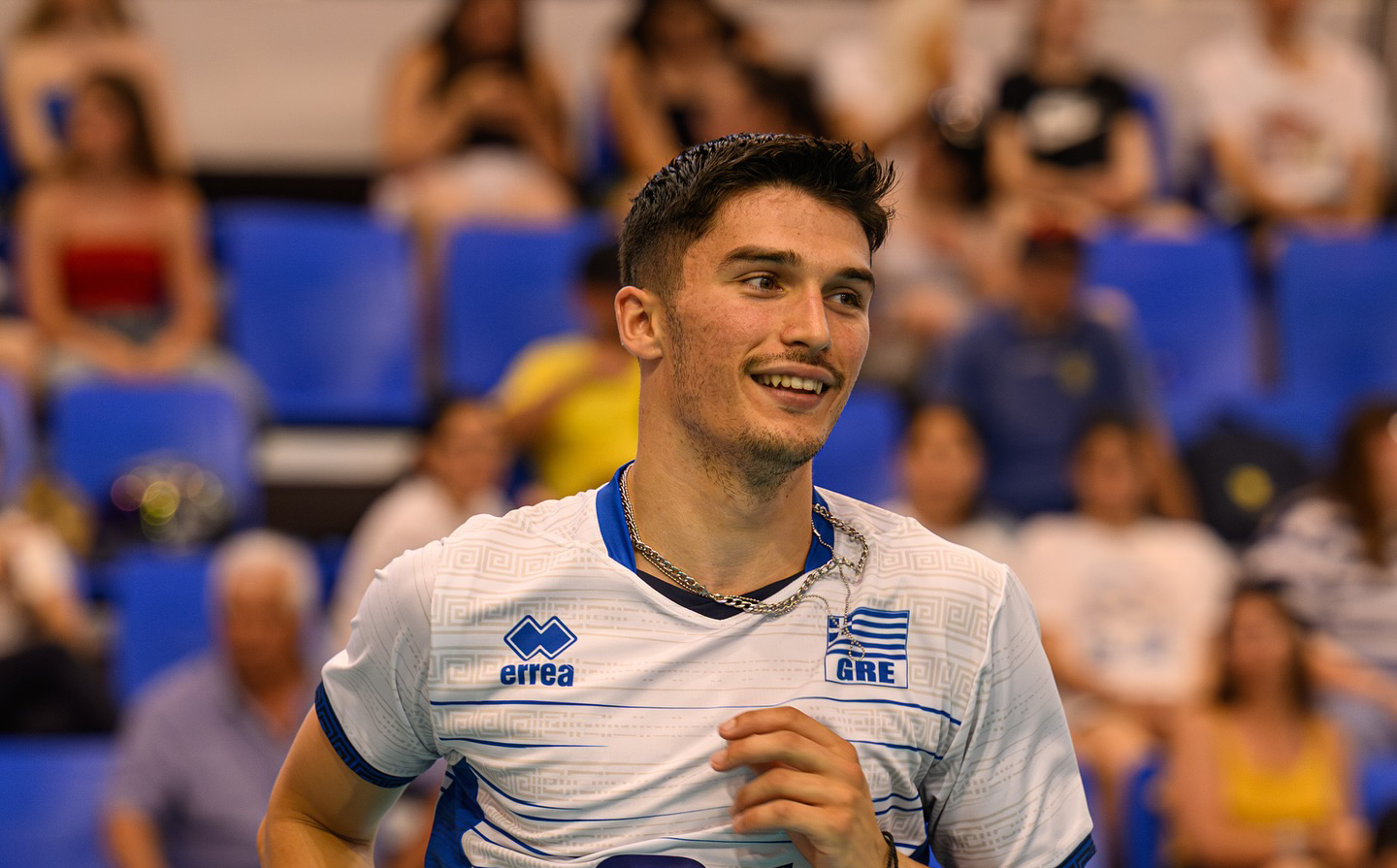 000 The Volley Lovers Smiling male athlete wearing a Greece jersey stands on a sports court with blue stadium seats in the background.
