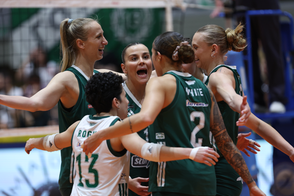 Female volleyball players in green uniforms celebrate together on the court, arms around each other after a point.