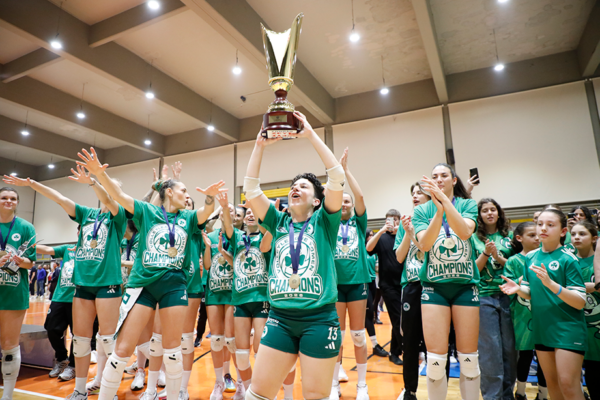 Girls volleyball team in green uniforms celebrate, lifting a large trophy overhead on an indoor court with cheering teammates nearby.