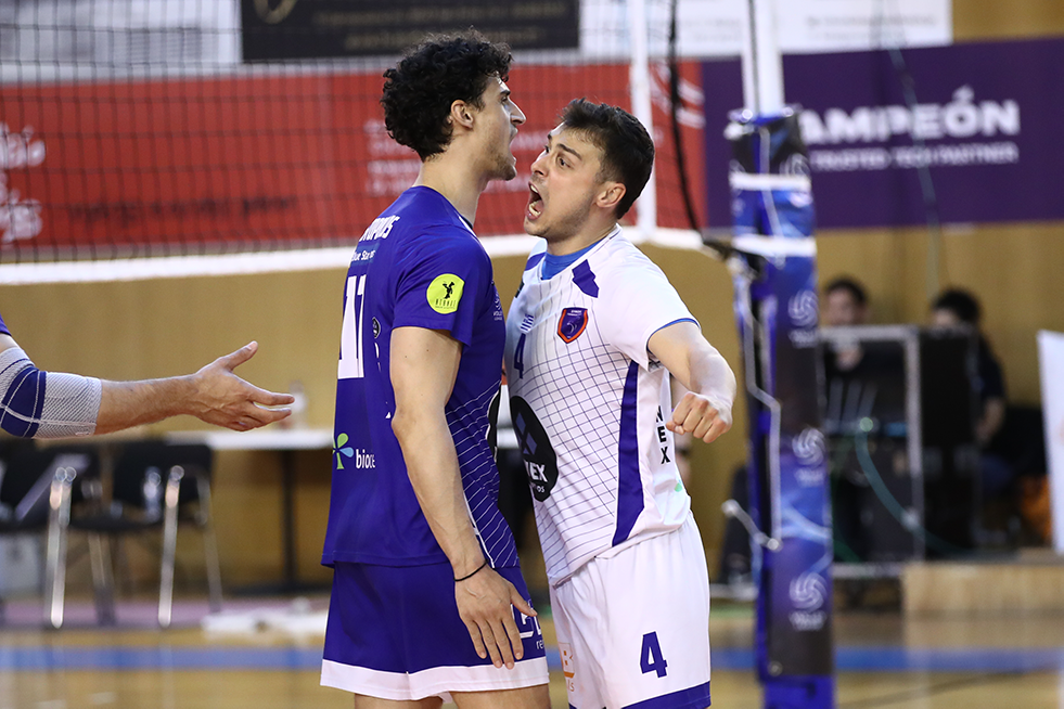 Two male volleyball players in blue and white jerseys confront each other inches apart near the net during a match, showing a tense moment.