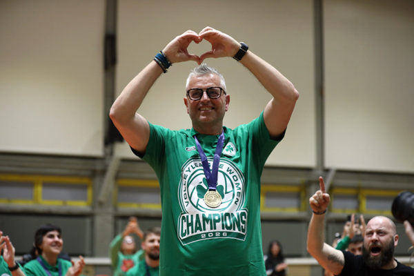 Man in green champions shirt with gold medal forming a heart shape with arms overhead, celebrating victory.