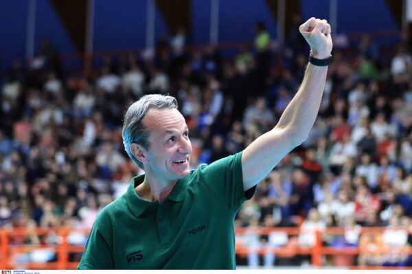Man in a green polo shirt raises a clenched fist in celebration at a crowded indoor sports arena in the background.