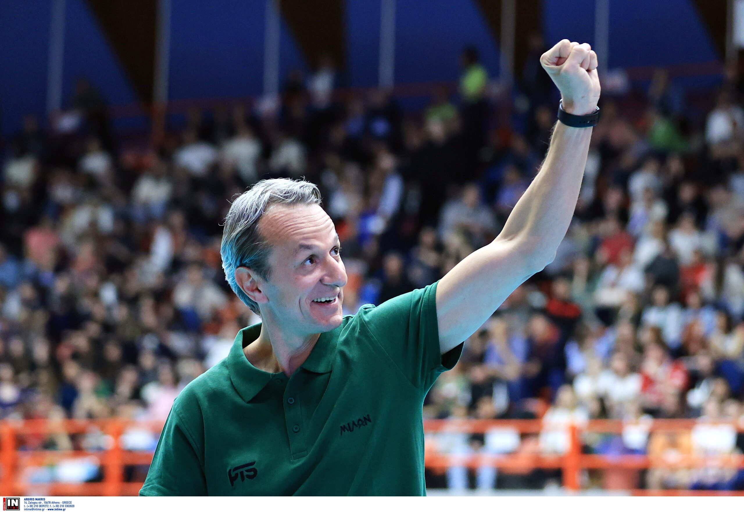 ÏËÕÌÐÉÁÊÏÓ ÌÉËÙÍ<br>ÔÅËÉÊÏÓ<br>ÖÁÉÍÁË ÖÏÑ ÊÕÐÅËËÏÕ 20232024 The Volley Lovers Man in a green polo shirt raises a clenched fist in celebration at a crowded indoor sports arena in the background.