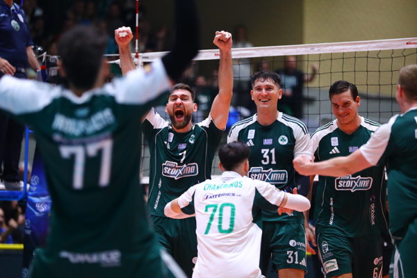 Volleyball players in green jerseys celebrate with raised arms at the net after scoring a point.