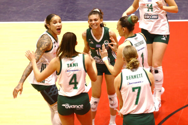 Six female volleyball players in green and white uniforms form a tight huddle on a gym floor, hands together in the center as they talk strategy.