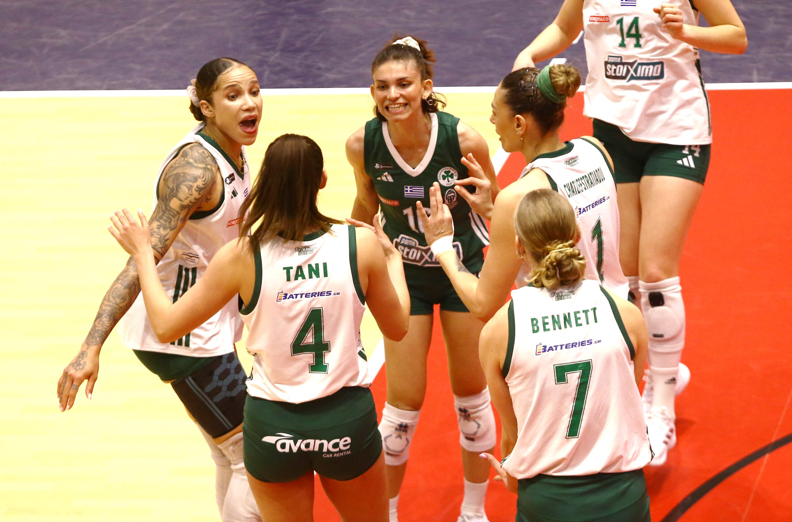 Six female volleyball players in green and white uniforms form a tight huddle on a gym floor, hands together in the center as they talk strategy.