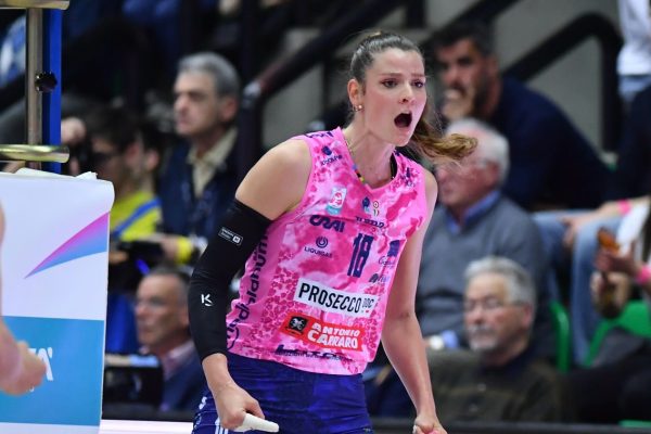 Female volleyball player in a pink jersey (#18) and blue shorts, shouting with mouth open during a match, spectators in the background.