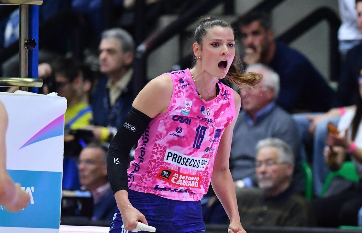 669830028185700563380619432201523894856516797n The Volley Lovers Female volleyball player in a pink jersey (#18) and blue shorts, shouting with mouth open during a match, spectators in the background.