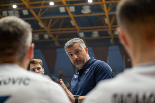 Coach with grey hair in a navy polo speaks to a group of athletes in a large indoor sports arena, players blurred in the foreground as they listen.