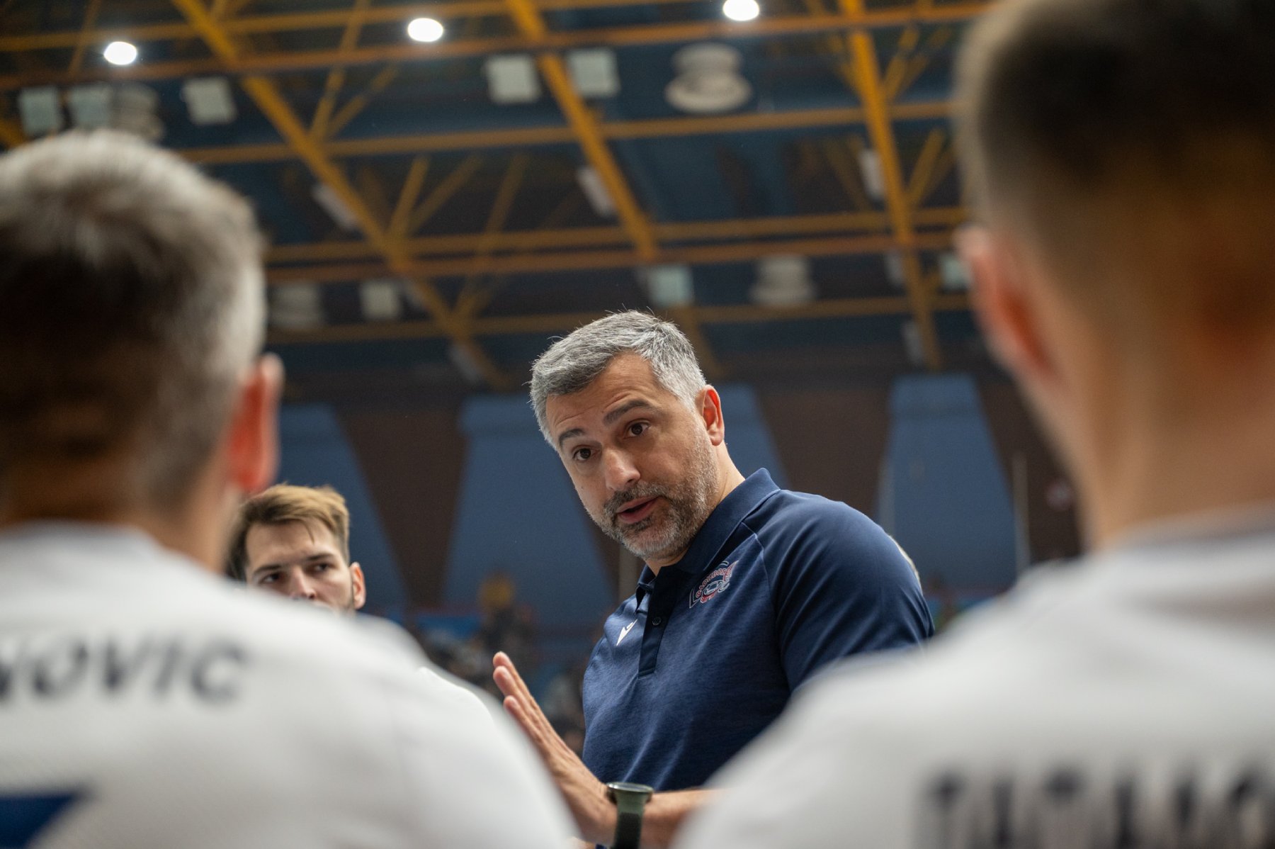 67243989814279701024658872922633163631549239n The Volley Lovers Coach with grey hair in a navy polo speaks to a group of athletes in a large indoor sports arena, players blurred in the foreground as they listen.