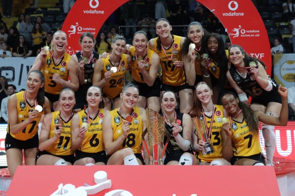 Women’s volleyball team in yellow and black uniforms cheerfully pose with medals and a large trophy after a championship win.