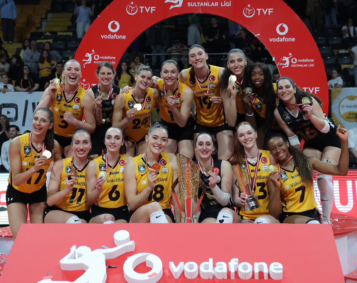673779743185755803310607435022447901073216912n The Volley Lovers Women’s volleyball team in yellow and black uniforms cheerfully pose with medals and a large trophy after a championship win.