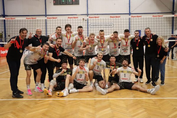 A sports team posing with medals and a trophy on an indoor court after a victory, players smiling and raising their arms in celebration.