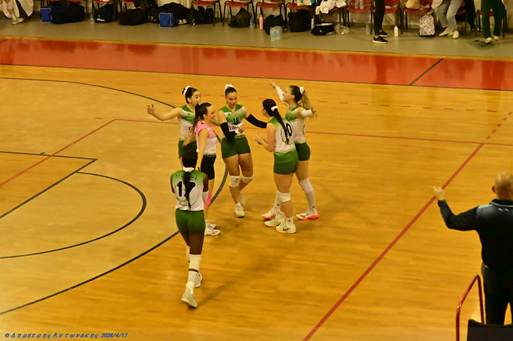 Women’s volleyball team in green and white uniforms celebrate a point on an indoor court, circling and high-fiving each other.