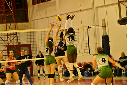 Volleyball match in progress as players leap at the net to block a ball while teammates watch from nearby court side.