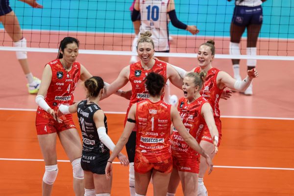 Group of female volleyball players in red uniforms huddle together on an orange court beside a net, celebrating a point.