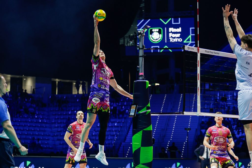 001 The Volley Lovers A volleyball player in a pink-purple uniform leaps near the net to hit the ball as others watch in a dark arena with blue seating.