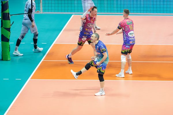 Volleyball players in pink uniforms celebrate on an orange court near the net with a teal boundary area nearby.