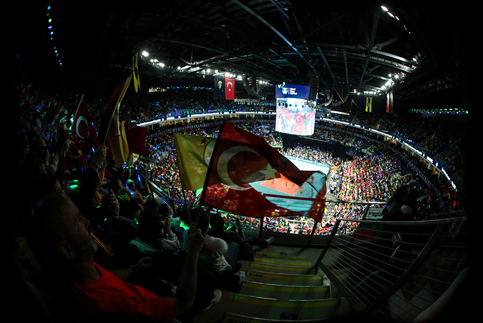 Crowd in a large indoor arena waving Turkish flags during a live event, with a circular court and big screen.