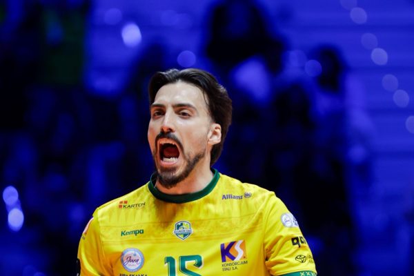Male volleyball player in yellow jersey number 15 shouting with mouth open, in a blue-lit arena background.