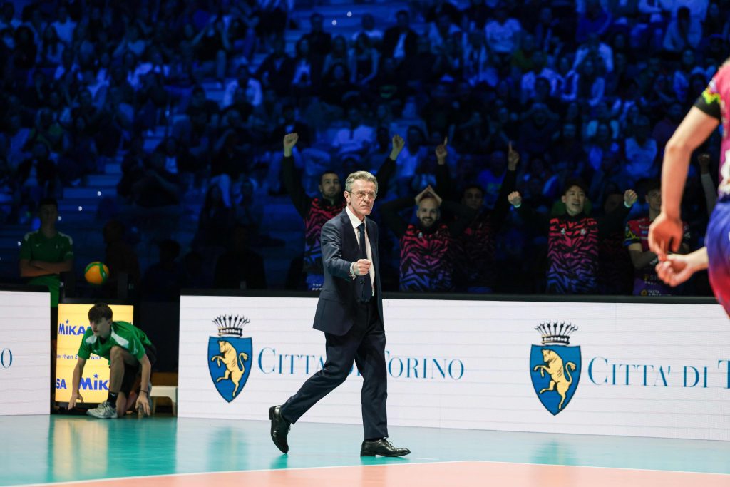 003 The Volley Lovers Man in a dark suit walks across an indoor volleyball court as a blue-lit crowd cheers in the stands behind him.