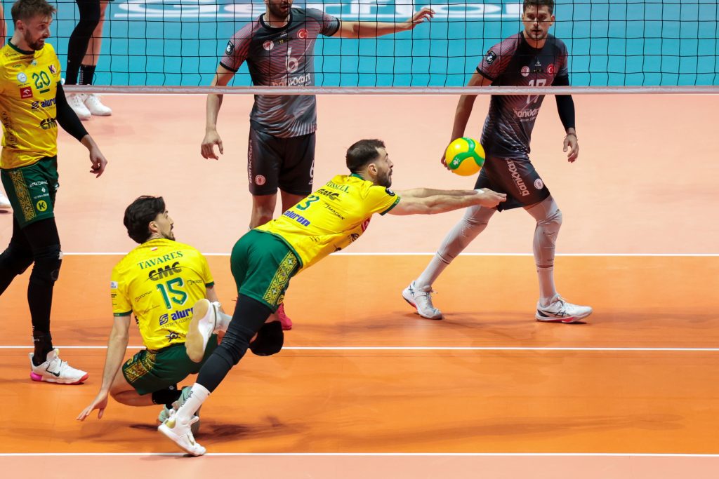 004 The Volley Lovers Volleyball players in yellow/green and gray uniforms reach for the ball near the net on an orange court during a match.