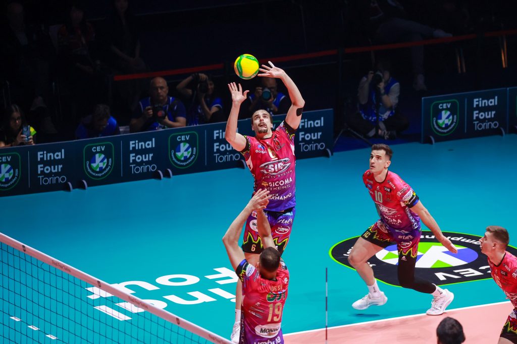 004 The Volley Lovers Volleyball players in red jerseys jump to set a bright ball above the net on a blue court with spectators in the background to the left and right.