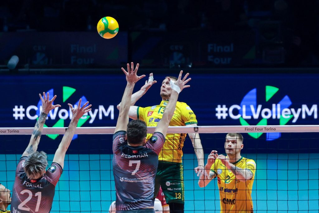 005 The Volley Lovers Volleyball players reach up at the net to block the ball during a match; yellow and dark uniforms visible in action moment.