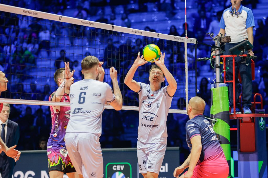 005 The Volley Lovers Volleyball players jump near the net as a bright yellow ball rises above, referee on right watching in a blue-lit arena.