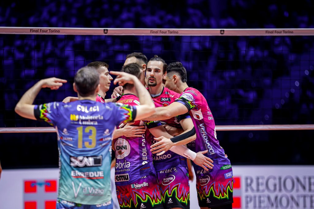 005 The Volley Lovers Group of volleyball players in pink and purple uniforms huddling at the net during a match on an indoor court.