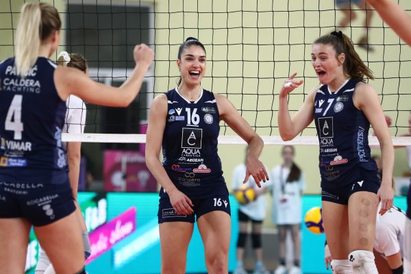 Three female volleyball players in navy uniforms celebrate at the net during a match, smiling and cheering.