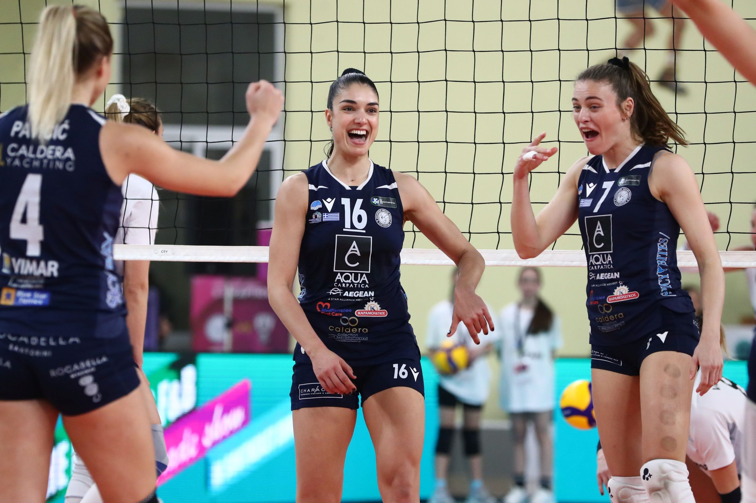 005072885 The Volley Lovers Three female volleyball players in navy uniforms celebrate at the net during a match, smiling and cheering.