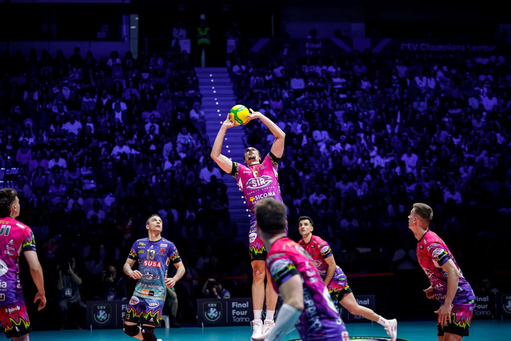 006 The Volley Lovers Volleyball player in pink jumps to spike the yellow ball, with a crowded arena in the background.