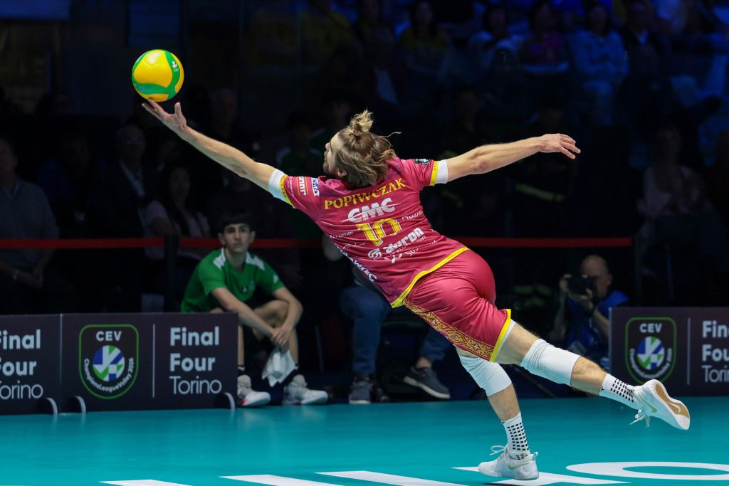 006 The Volley Lovers Volleyball player in a pink uniform leaps to strike a yellow ball on an indoor court with spectators in the darkened stands behind her.