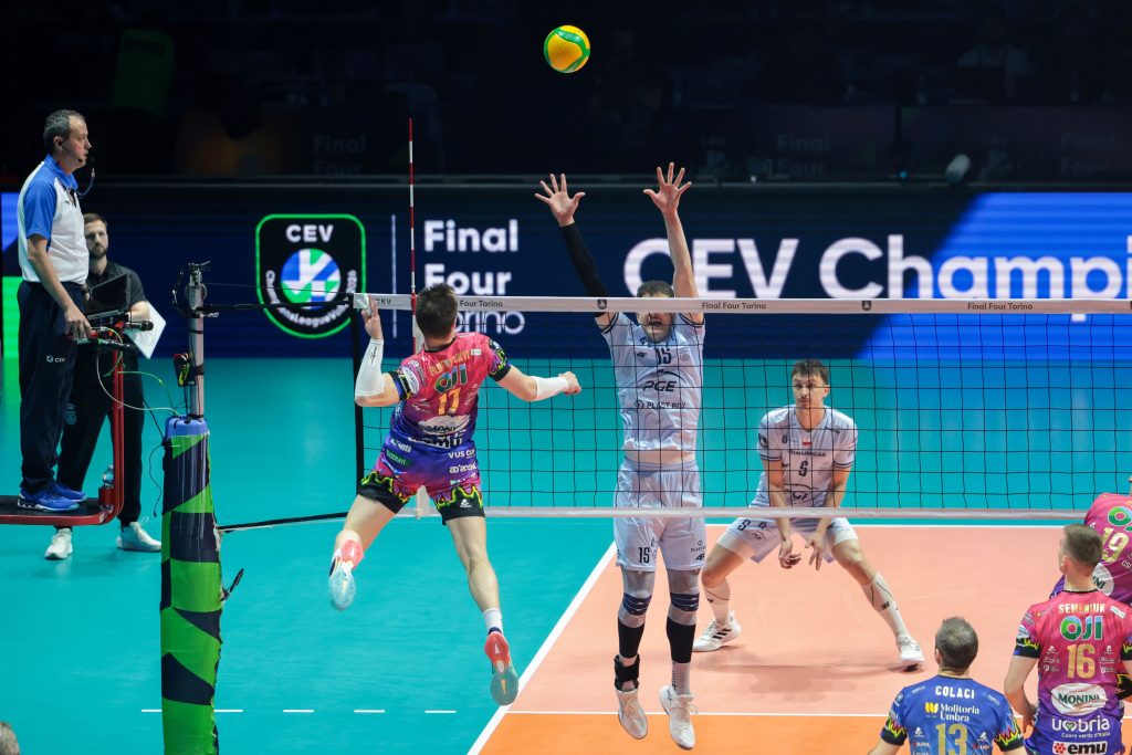 008 The Volley Lovers Volleyball action at the net: a pink-clad spiker jumps to hit while white-uniform players raise hands to block; referee watches from left.