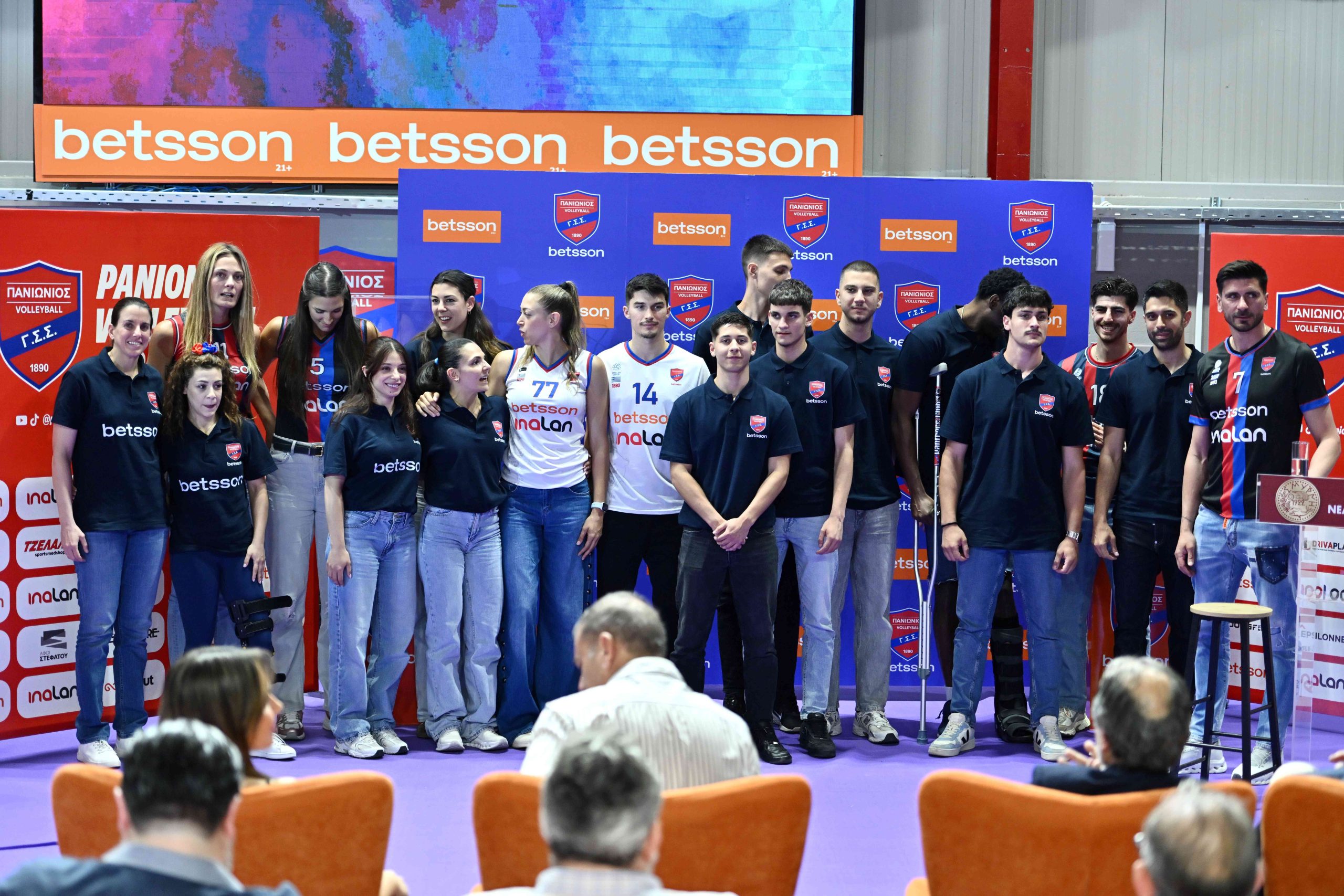 01 The Volley Lovers Group of volleyball players and staff posing for a photo on a stage with sponsor banners in the background.