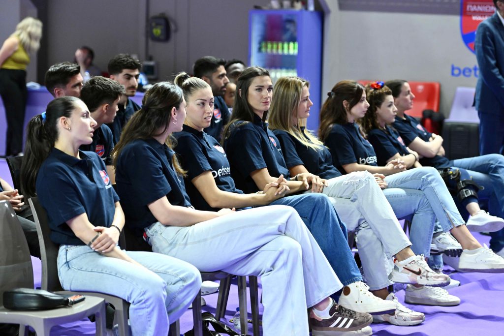 04 The Volley Lovers Row of people in navy shirts seated in a line at an event, facing forward on a purple-floored venue.