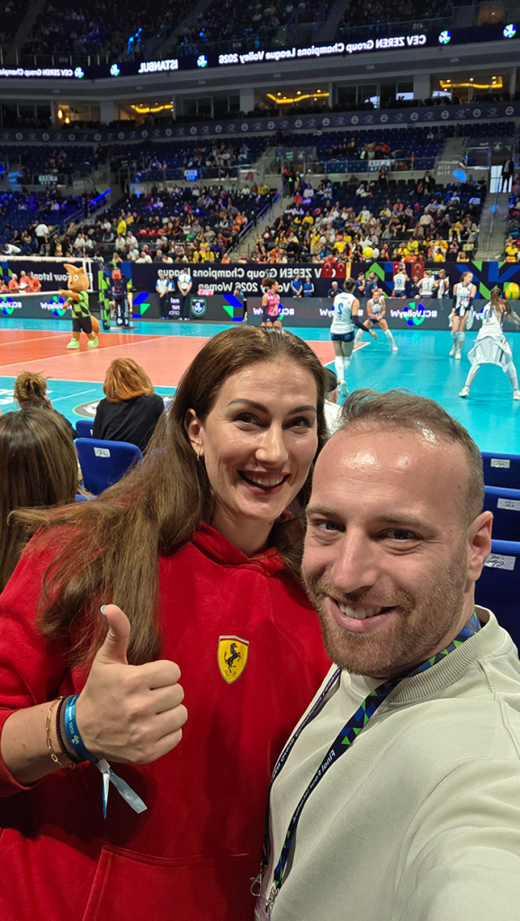 Two people take a cheerful selfie courtside at an indoor volleyball match; woman in red Ferrari hoodie shows a thumbs up, man smiling beside her, with players and a crowd in the background.