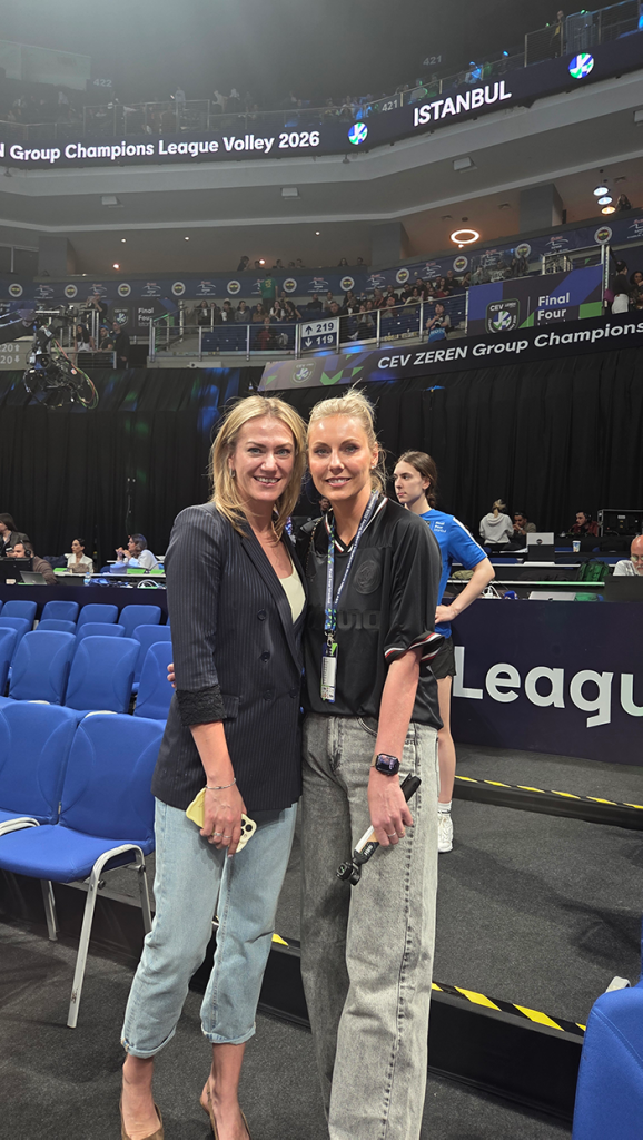 Two women pose for a photo in a volleyball arena, smiling, with blue seats and spectators behind them.