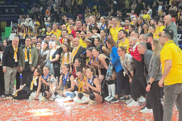 Volleyball team posing with a large trophy on the court as fans cheer behind them.