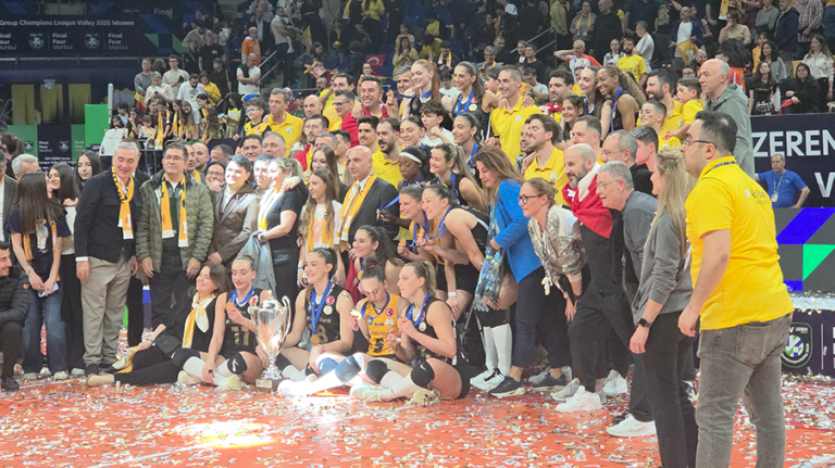 20260503224312 The Volley Lovers Volleyball team posing with a large trophy on the court as fans cheer behind them.