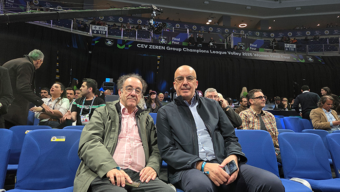Two men seated in blue arena chairs at a volleyball event, with spectators around them and a banner overhead mentioning the CEV Champions League Final Four.