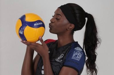 Female volleyball player in a dark jersey kissing a Mikasa volleyball against a light background.",