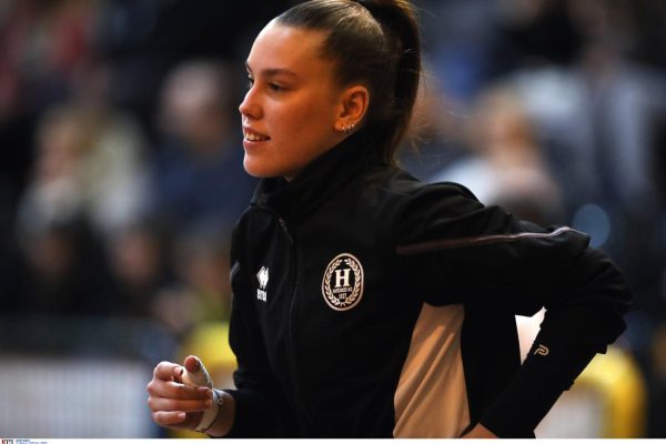Female athlete in a black and beige track jacket running, hair in a ponytail, blurred crowd in the background.