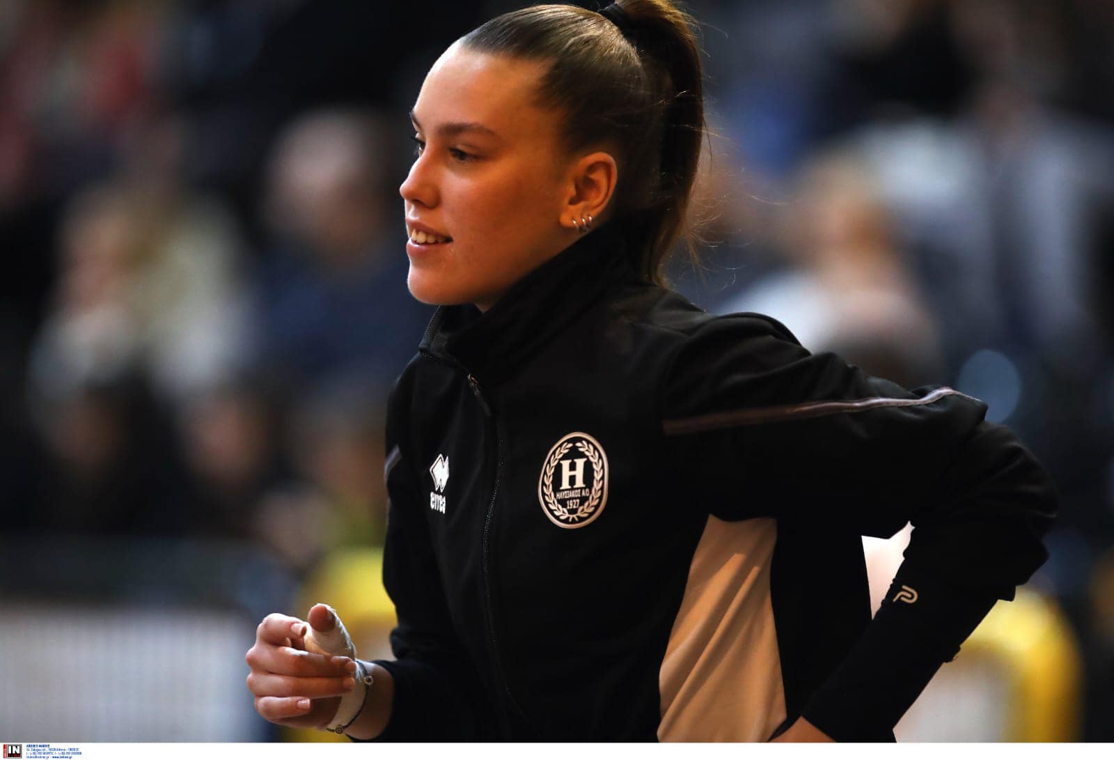 48235372420293582375620176360159759133973674n The Volley Lovers Female athlete in a black and beige track jacket running, hair in a ponytail, blurred crowd in the background.