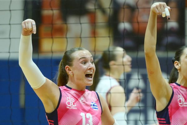Two female volleyball players in pink jerseys celebrate with arms raised behind a net in a gymnasium.