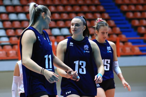 Three female volleyball players walk together on court, wearing blue uniforms with numbers 11, 13, and 15, in a gym with orange seats behind them.