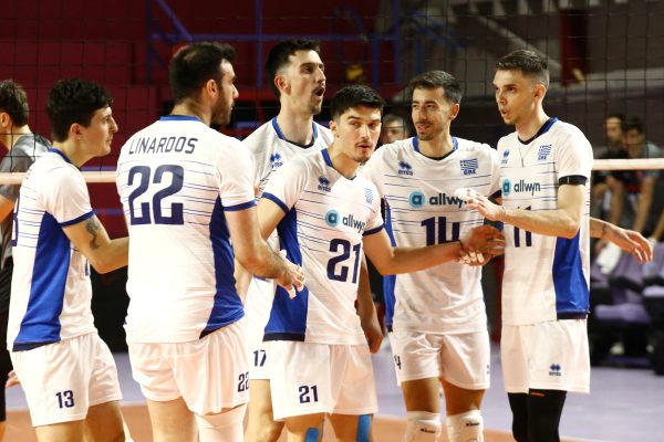Group of male volleyball players in white and blue uniforms huddling on an indoor court near the net.